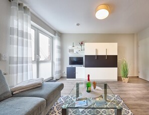 Una sala moderna con un sofá, una mesa de centro de vidrio y una zona de cocina con armarios blancos y un televisor.