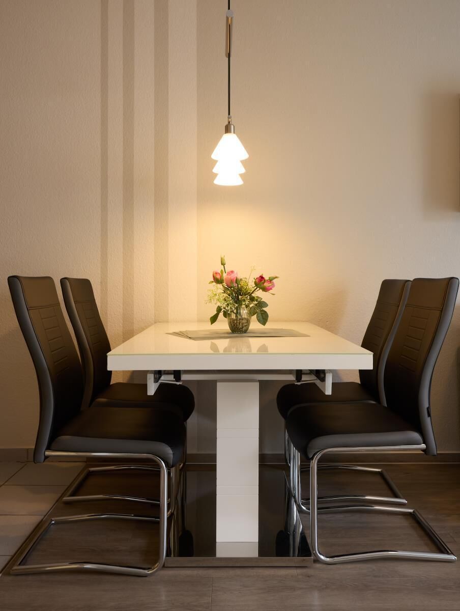 Spiseplads med et hvidt bord, fire stole, en hængende lysekrone og en vase med blomster.