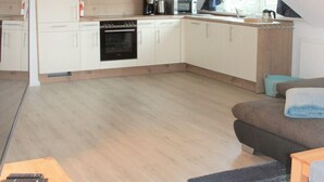 A compact kitchen with white cabinets, a microwave, and an oven. There is a wooden table with a bowl and a remote control. A blue rug is on the floor, and a brown couch is in the living area. - Villa Döser Strand penthouse apartment 11 (Cuxhaven)