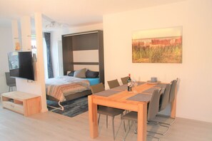 A modern bedroom with a bed, bedside table, and a TV. There is a dining area with a table, chairs, and a painting on the wall. - Villa Döser Strand, Penthouse Apartment 11 (Cuxhaven)