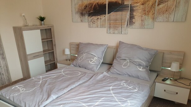 A neatly made bed with a grey bedspread and pillows, a bedside table with a lamp, and a small cabinet to the left.