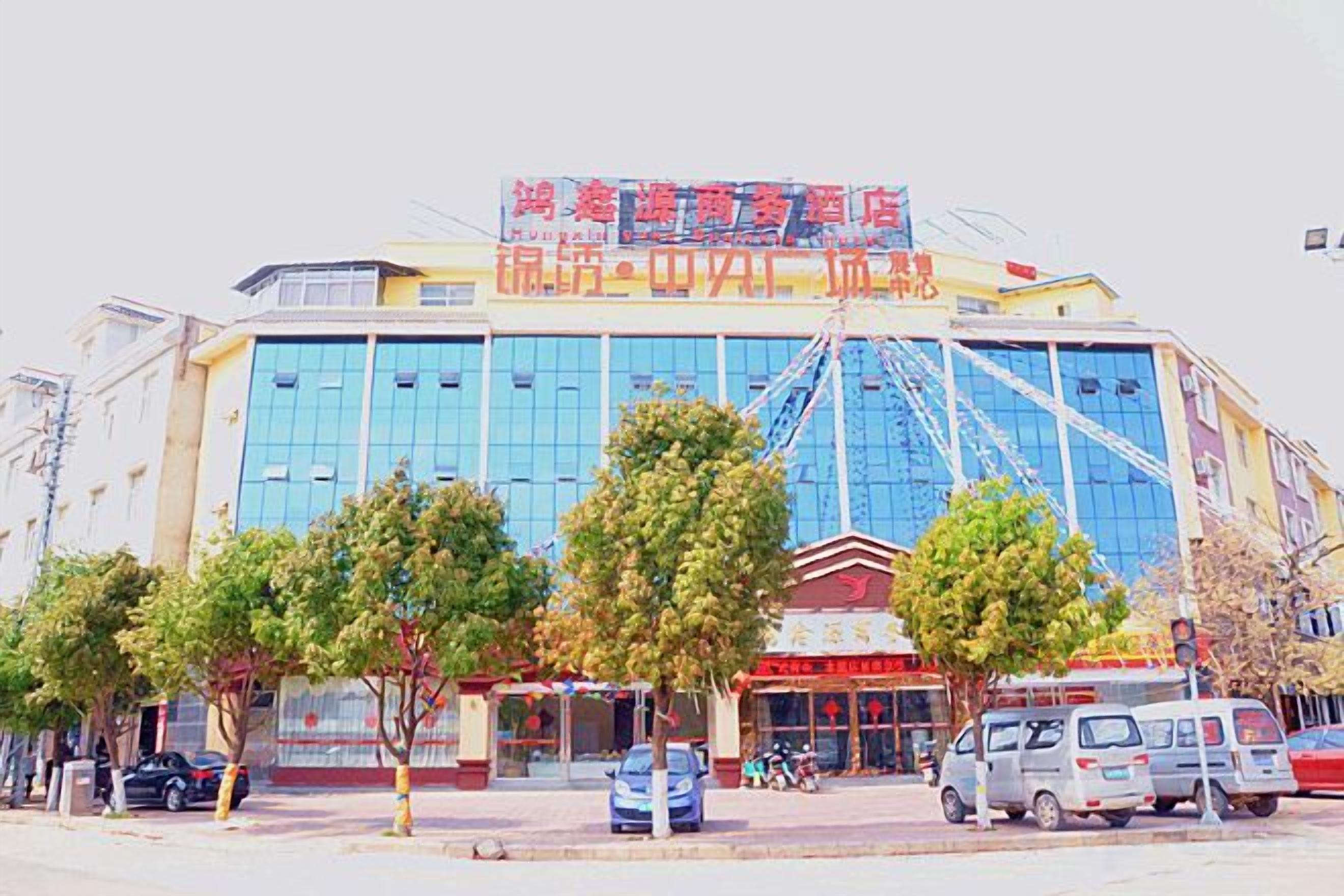 A multi-story building with a prominent sign, several trees in front, and parked vehicles.