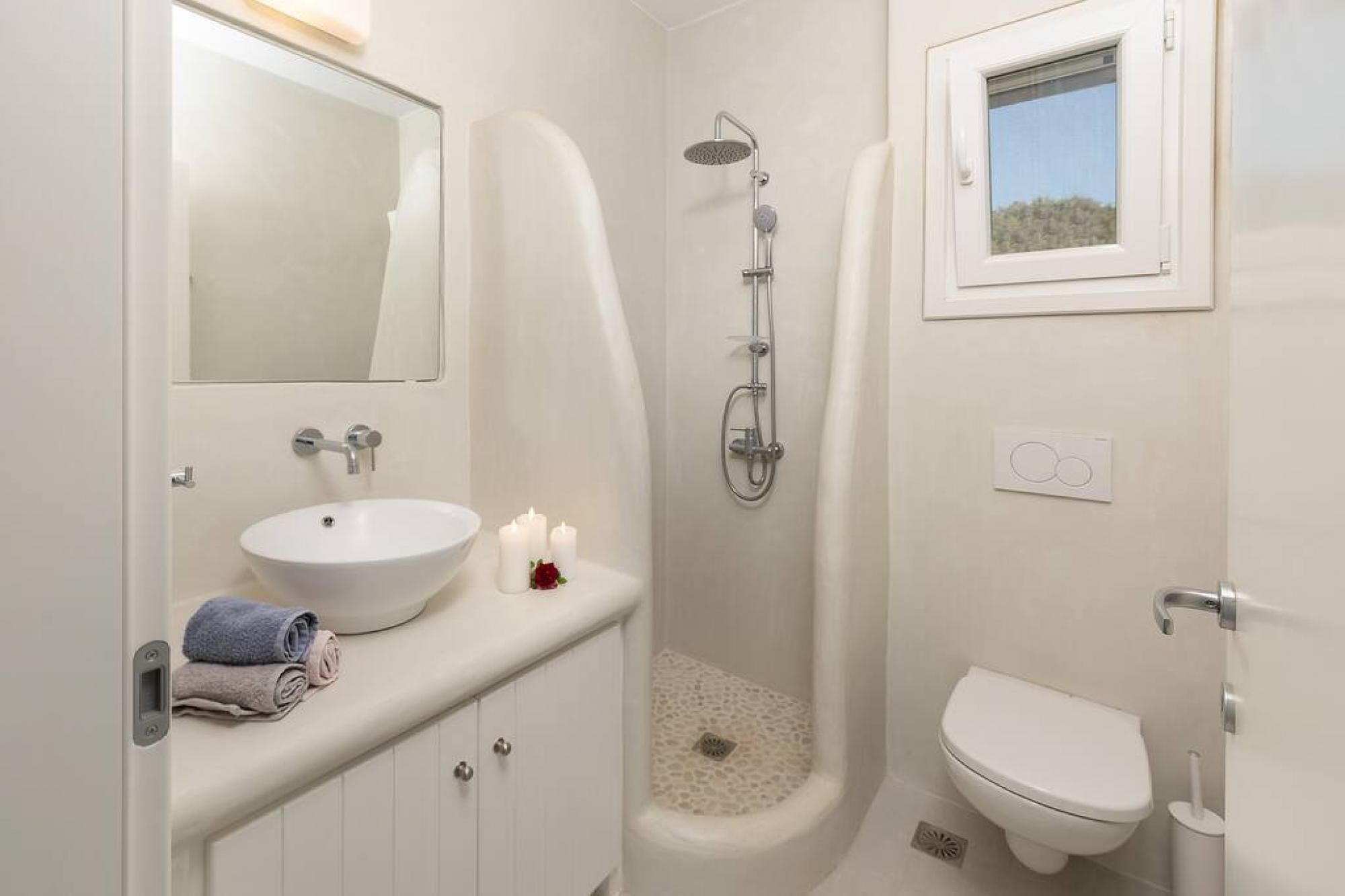 A bathroom with a white sink, mirror, and toilet. There is a shower with a handheld showerhead and a window above the toilet.