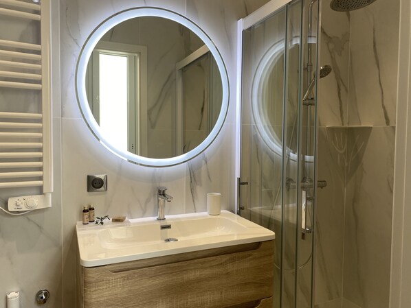 A bathroom with a round mirror, a sink, and a glass-enclosed shower. - Nancy Thermal Blandan : Suite Stanislas (Nancy)