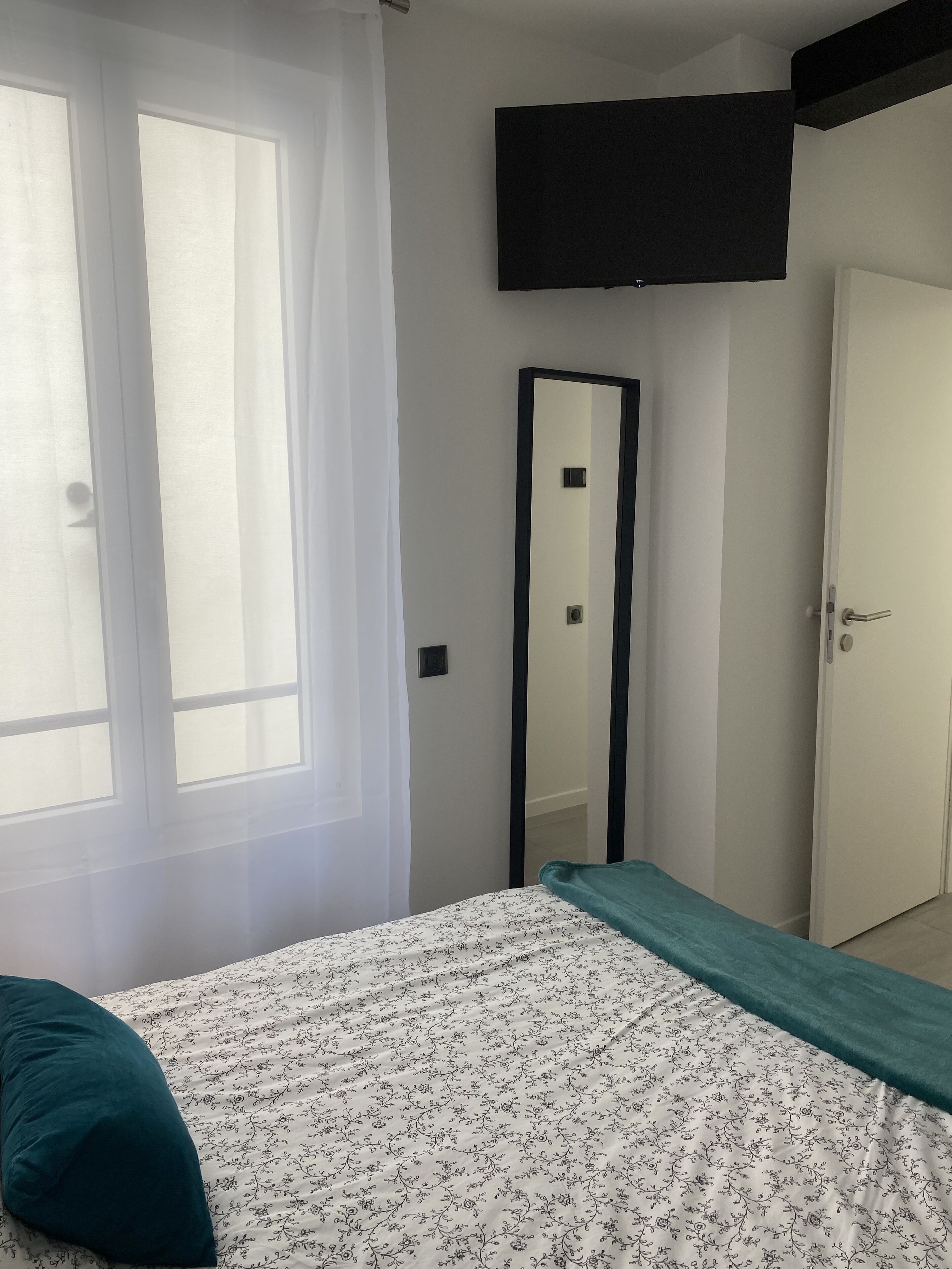 A bedroom with a bed, a mirror, a door, and a window with sheer curtains.
