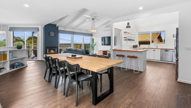 A spacious living area with a dining table, chairs, and a fireplace. There is a kitchen with a bar area and a large window. - Dune Views, Gas Fire and Pet friendly. (Ocean Grove)