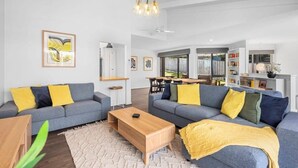 A modern living room with two grey sofas, yellow cushions, a wooden coffee table, and a kitchen in the background. - Dune Views, Gas Fire and Pet friendly. (Ocean Grove)