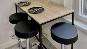 Un petit coin repas avec une table en bois, des tabourets noirs, une horloge murale et une petite étagère sur laquelle est posée une plante.