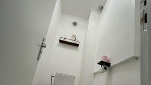 Une petite salle de bain blanche, avec des toilettes, une étagère sur laquelle est posée une petite plante, et une fenêtre ronde.