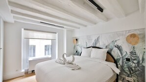 A bedroom with a large bed, white bedding, a window with blinds, a wall with a leafy pattern, and a ceiling with exposed beams. - 11.Flat 8 person St Lazare Galerie LaFayette 8ème (Paris)