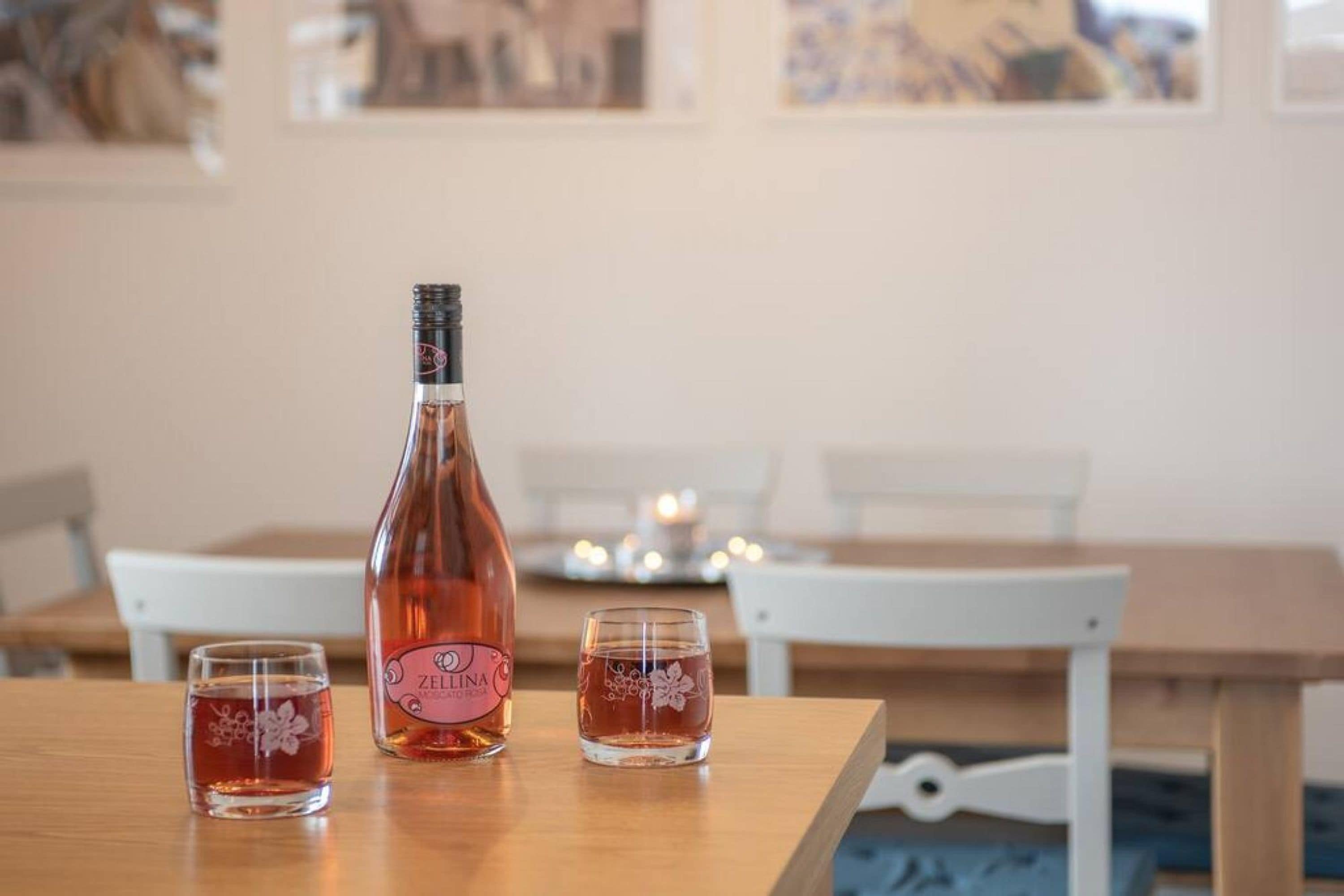 Une bouteille de vin Zelkina et deux verres sur une table en bois, dans un coin repas avec des chaises blanches et une bougie en arrière-plan.