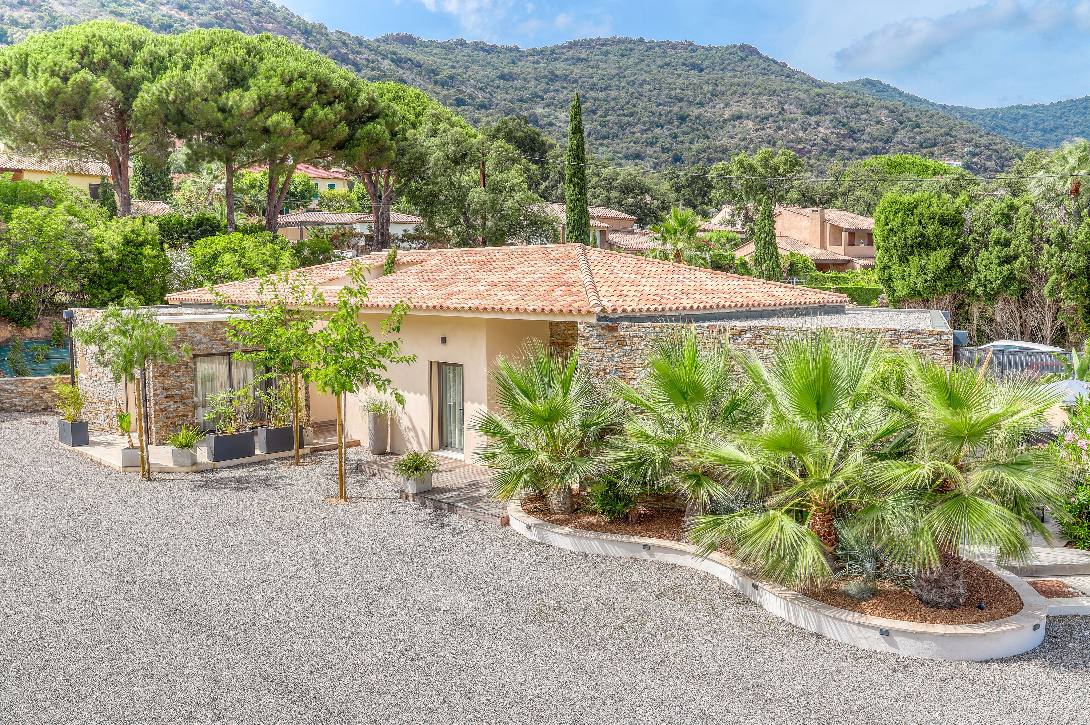 A villa with a tiled roof, surrounded by a gravel driveway and lush greenery, including palm trees and potted plants.