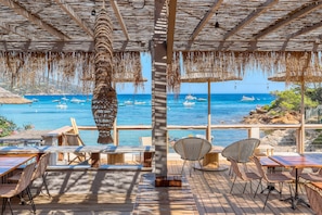 Een terras met tafels en stoelen met uitzicht op een heldere blauwe zee met boten.