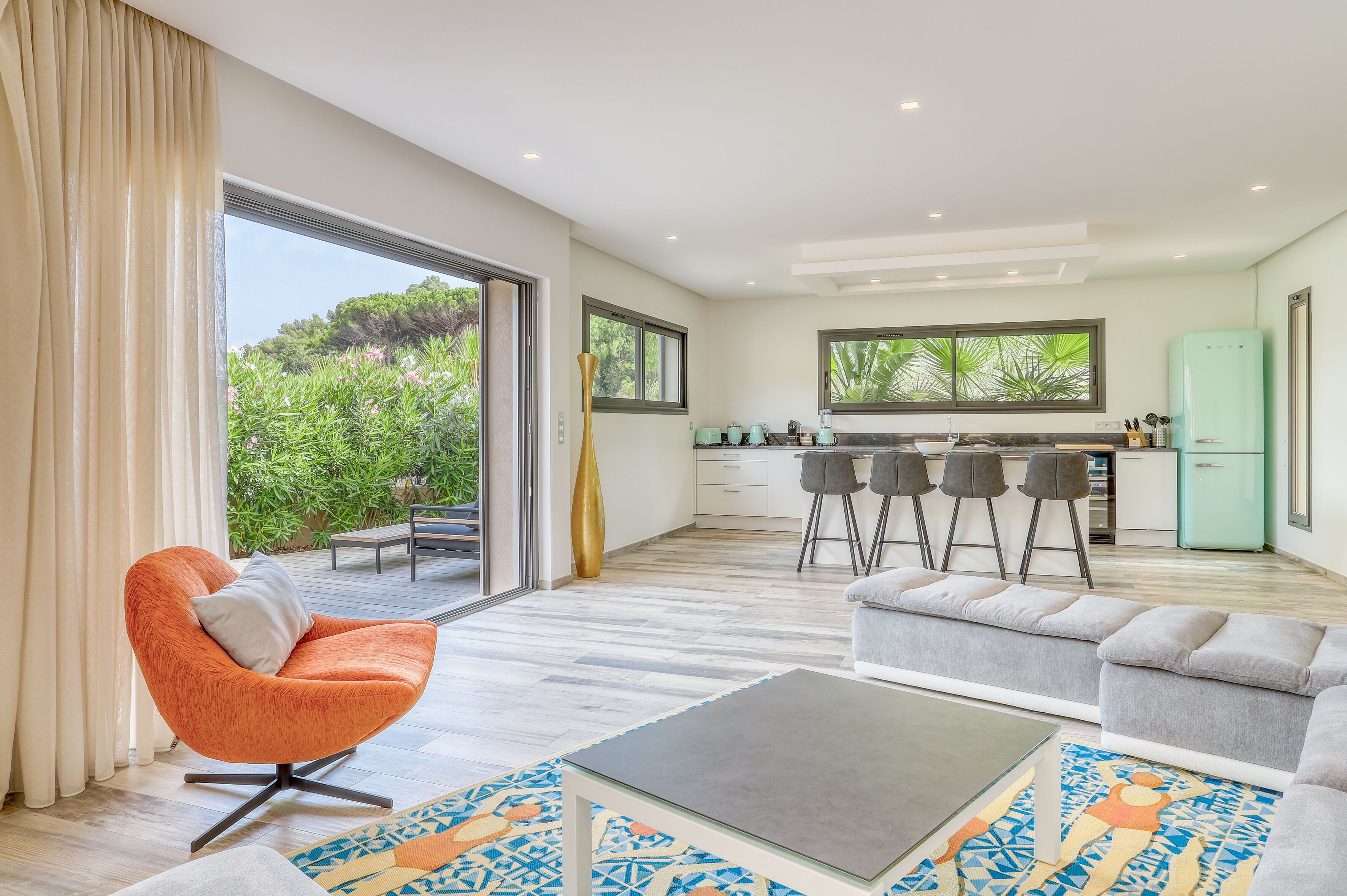 A modern living room with a sofa, a coffee table, and a chair. There is a kitchen area with a fridge and bar stools. Large windows provide a view of greenery outside.