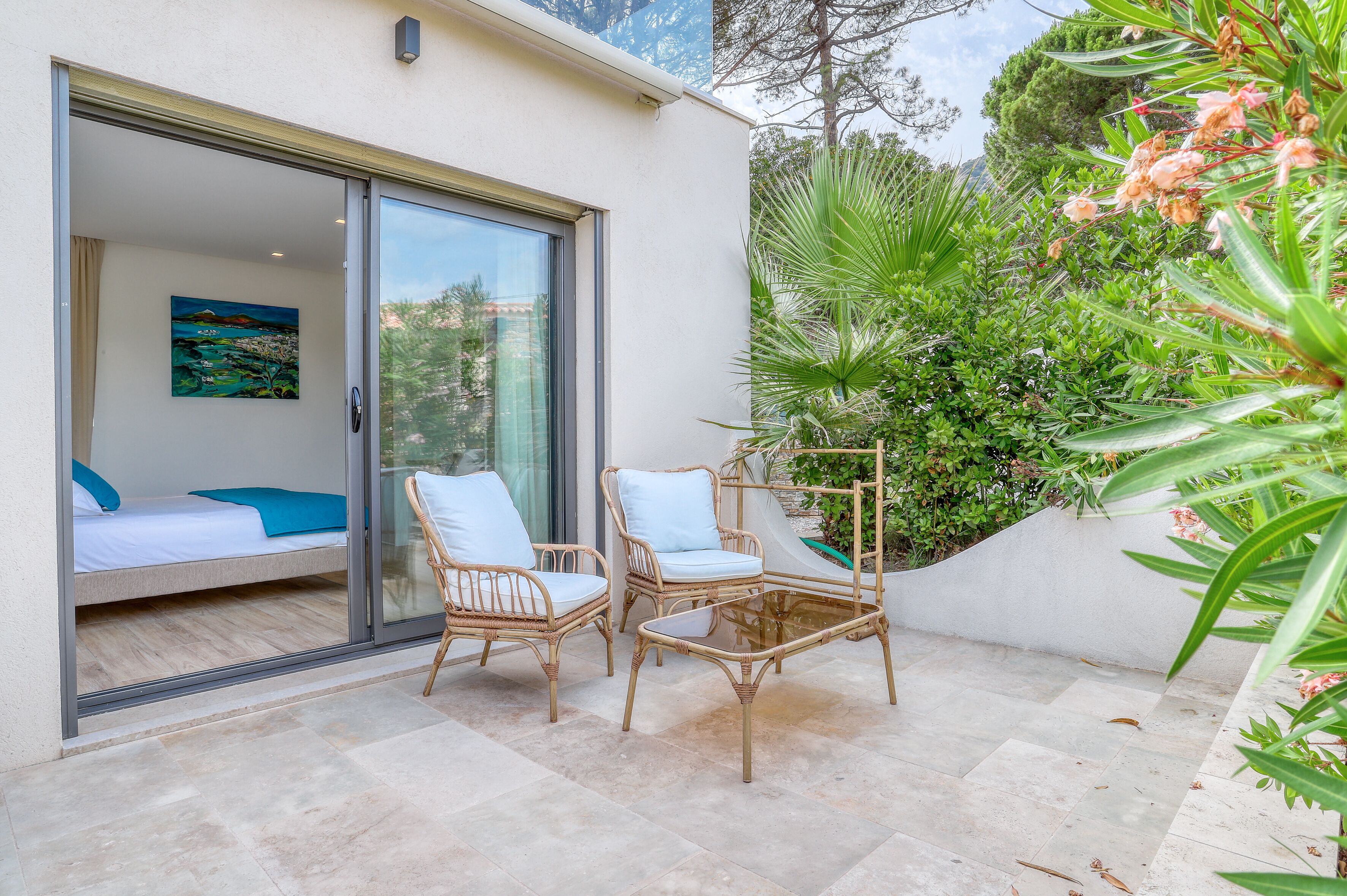 A modern outdoor patio with wicker furniture, a glass door leading to a bedroom with a bed and a painting, and lush greenery.