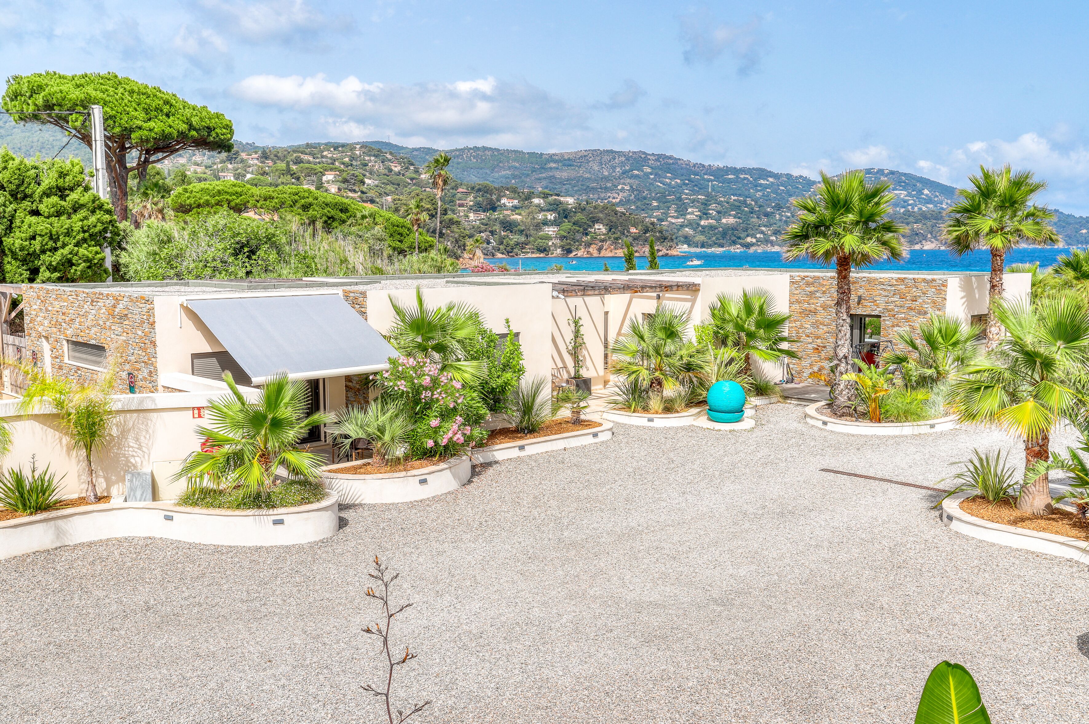 A rooftop terrace with a gravel ground, surrounded by palm trees and a view of the sea and hills.