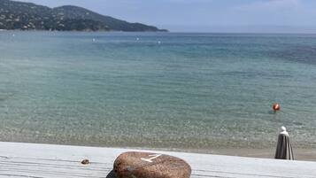 A table with a glass of a beverage, a stone, and an umbrella, overlooking a clear sea with a distant shoreline and hills.