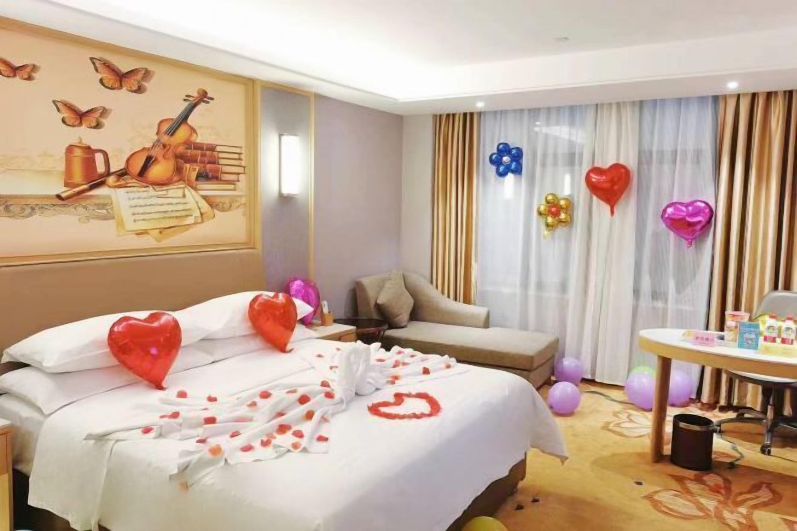Ein Hotelzimmer mit Bett, Schreibtisch und Stuhl. Auf dem Bett liegen Ballons und herzförmige Dekorationen.