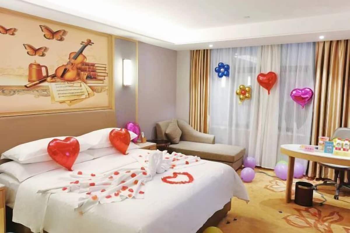 A hotel room with a bed, a desk, and a chair. There are balloons and heart-shaped decorations on the bed.
