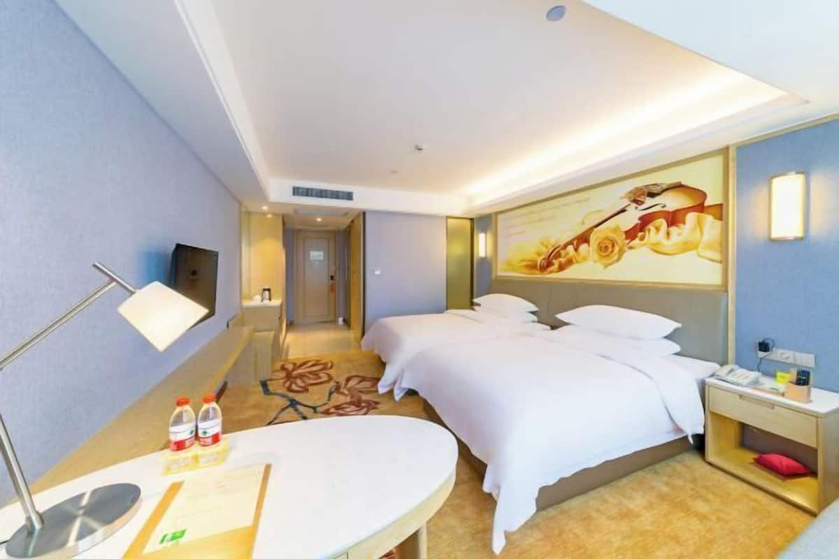A hotel room with two beds, a desk, a chair, a flat-screen TV, and a wall-mounted lamp.
