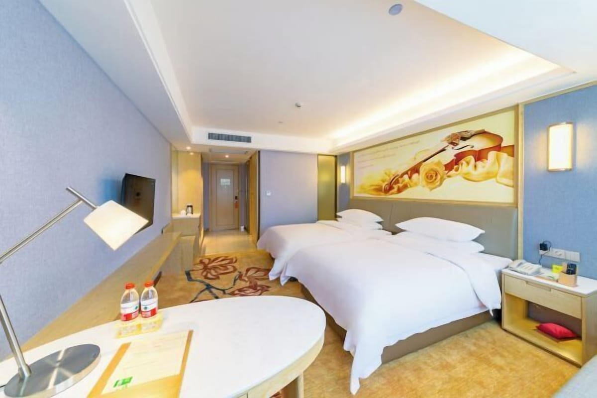 A hotel room with two beds, a desk with a lamp, a small table, and a wall-mounted TV.