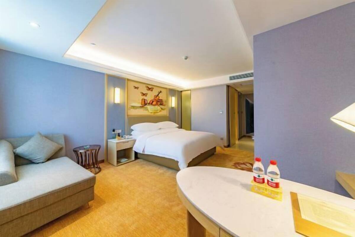 A modern hotel room with a sofa, a bed, a bedside table, a mirror, and a small table with bottles.