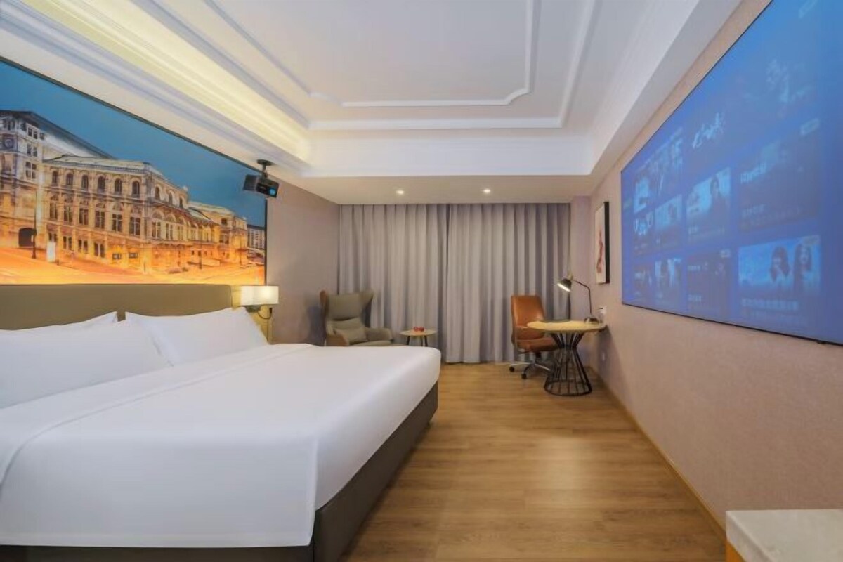Habitación de hotel con una cama grande, un escritorio con silla, una pantalla de proyector y un mural de un edificio.