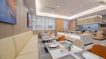 A modern dining area with a long table, cushioned seating, and a kitchen in the background.