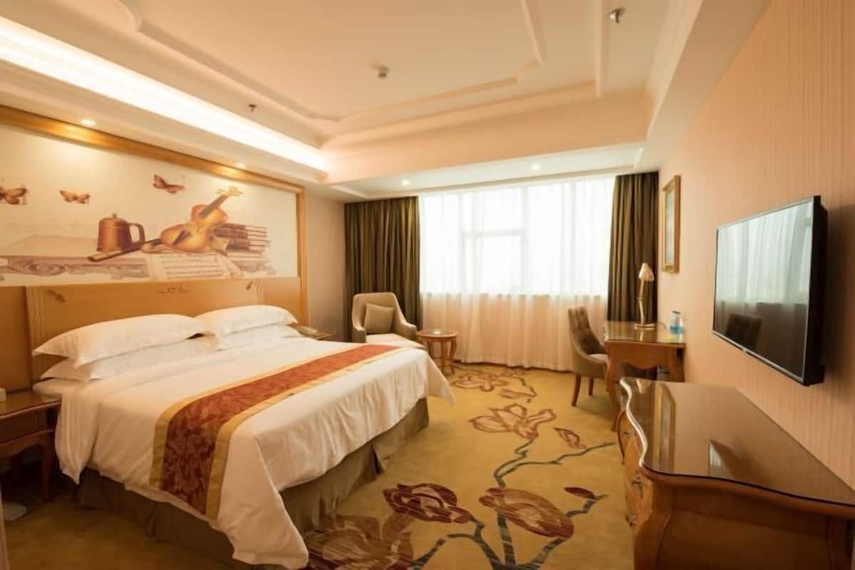 Une chambre d’hôtel avec un grand lit, un bureau, une chaise, une télévision et une fenêtre avec des rideaux.