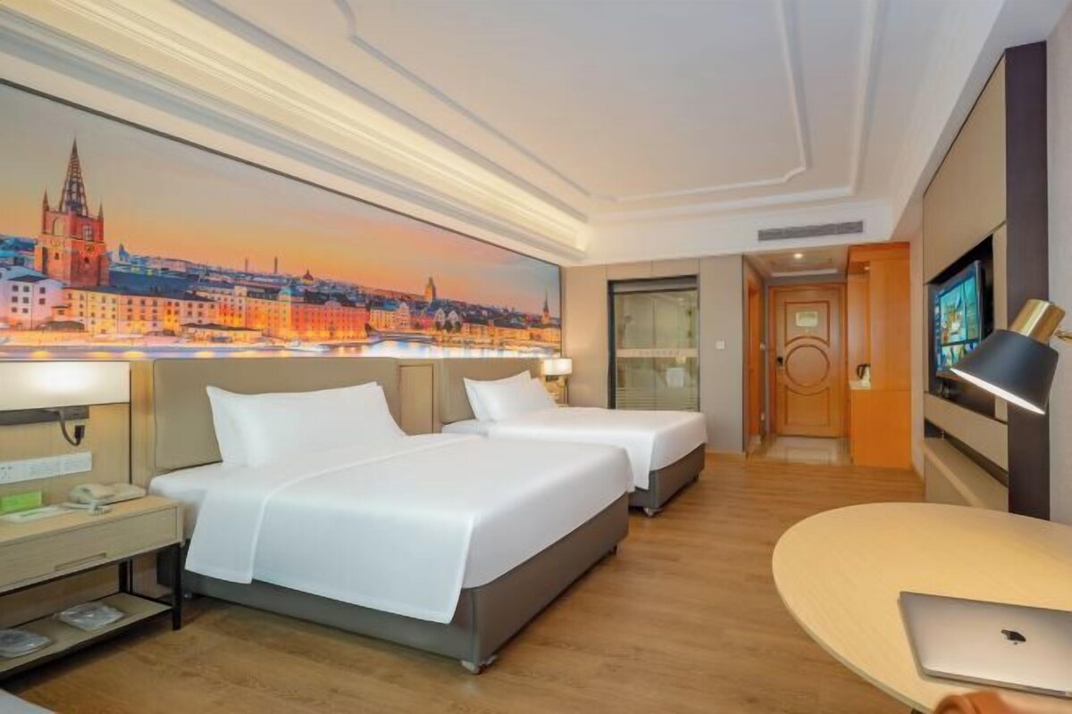 A hotel room with two beds, a desk with a lamp, a laptop on a table, and a cityscape painting on the wall.