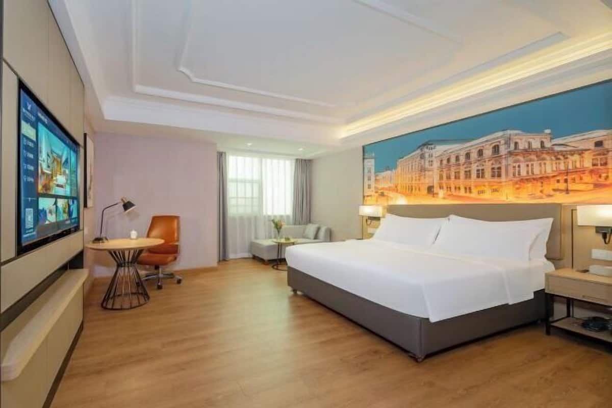 Une chambre d’hôtel avec un grand lit, un bureau avec un ordinateur, une chaise, une petite table et un tableau représentant un bâtiment accroché au mur.