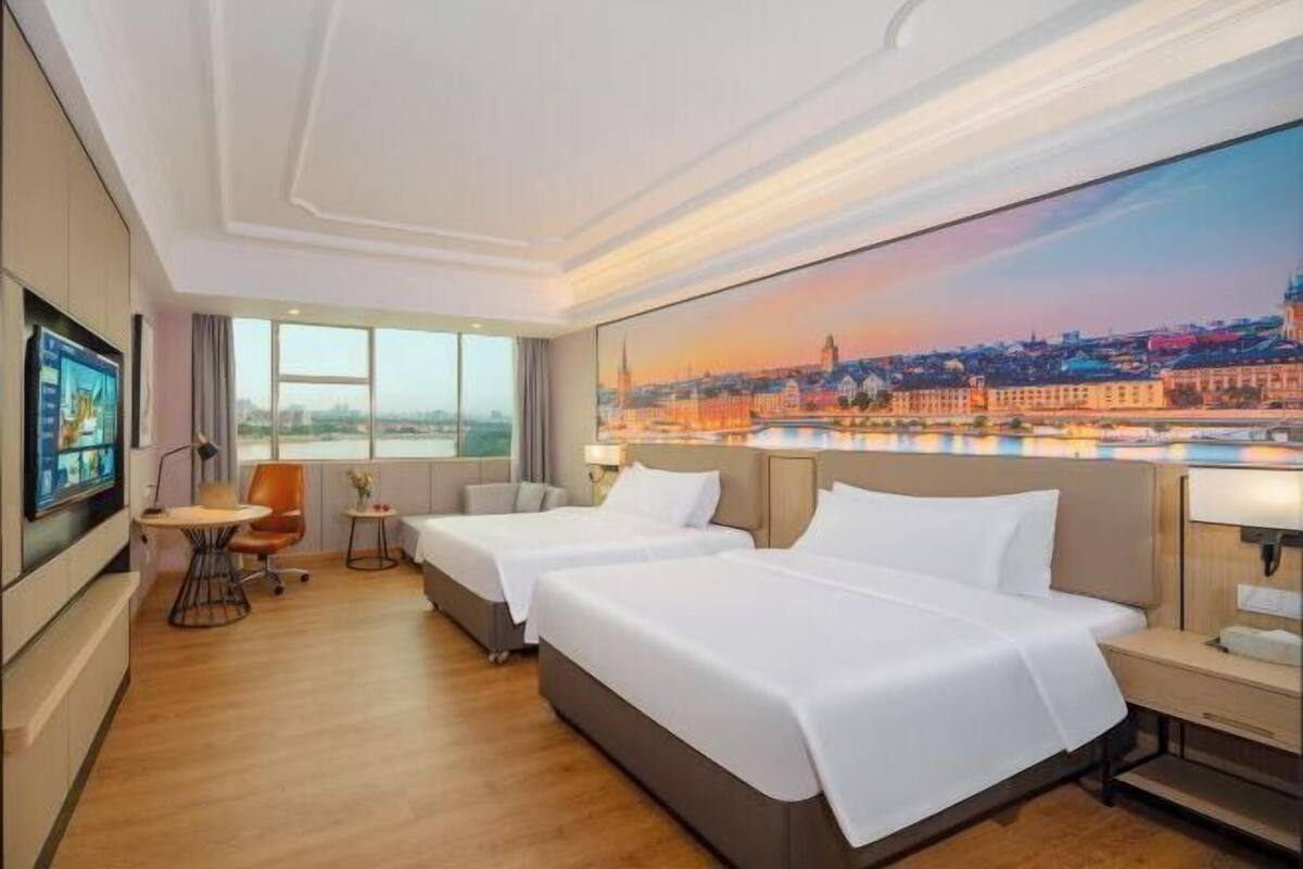 Quarto de hotel com duas camas, televisão, uma mesinha e um quadro de uma paisagem urbana.