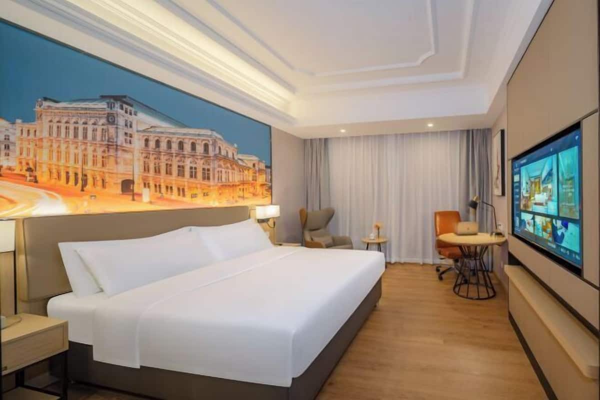 Une chambre d’hôtel avec un grand lit, un bureau, une chaise, une télévision et un tableau représentant un bâtiment accroché au mur.