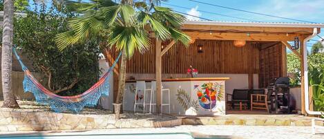 Zona junto a la piscina con bar de madera, asientos exteriores y una hamaca.