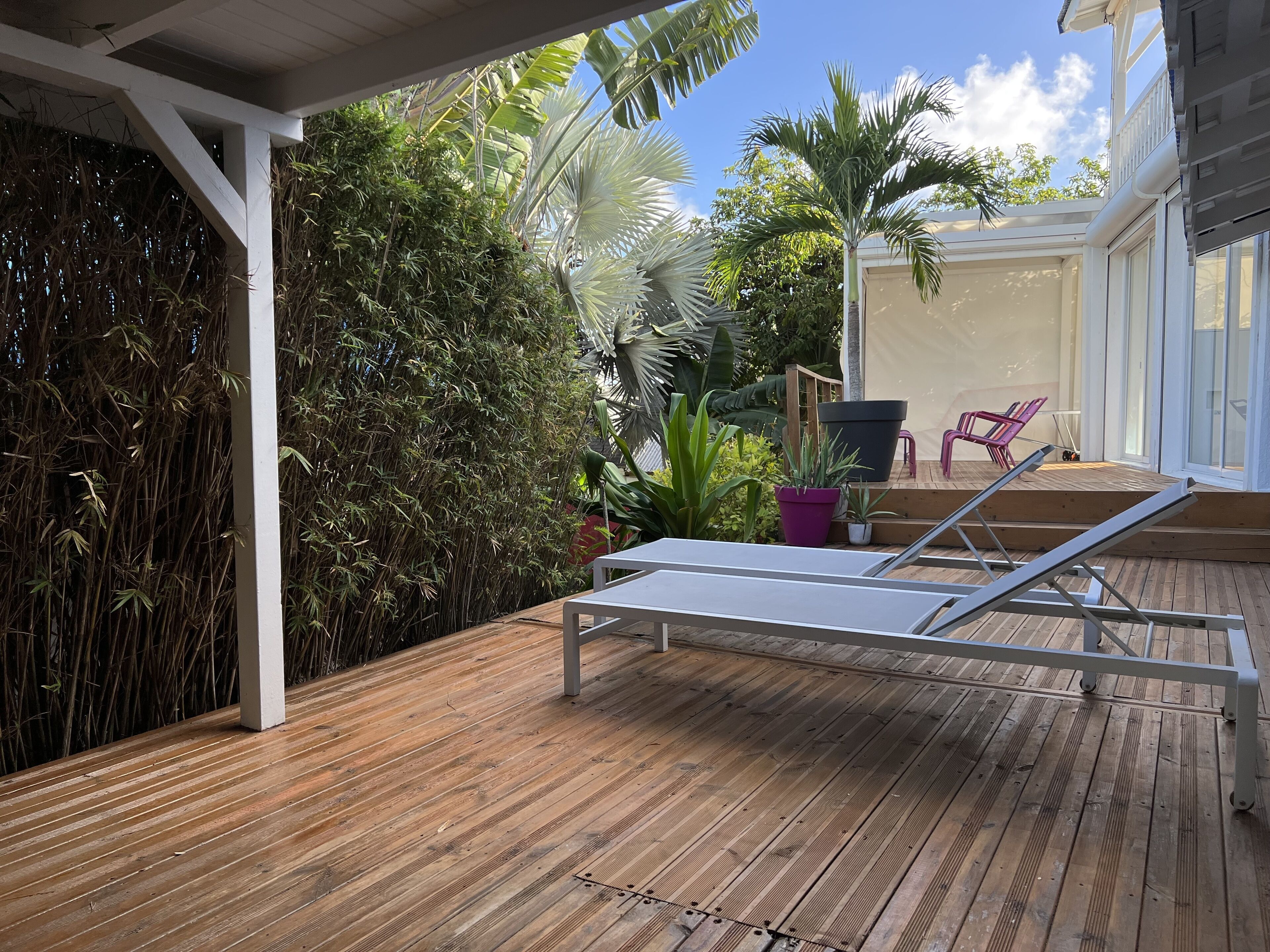 A wooden deck with a metal sun lounger, surrounded by lush greenery and a white pergola.