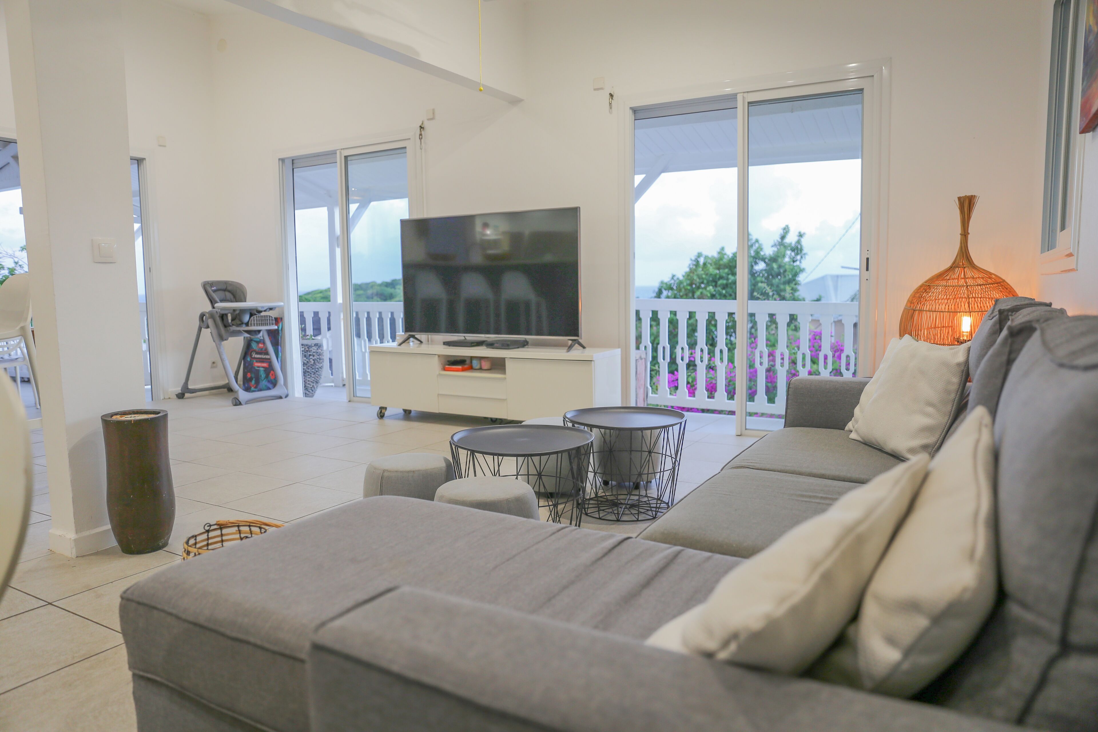 En moderne stue med en grå sofa, et fladskærms-tv, to runde borde og et stort vindue med en balkon.