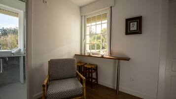 Una habitación con piso de madera, un sillón estampado, una ventana con vista y un taburete pequeño.
