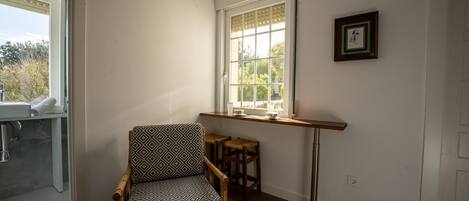 A room with a wooden floor, a patterned chair, a window with a view, and a small stool.