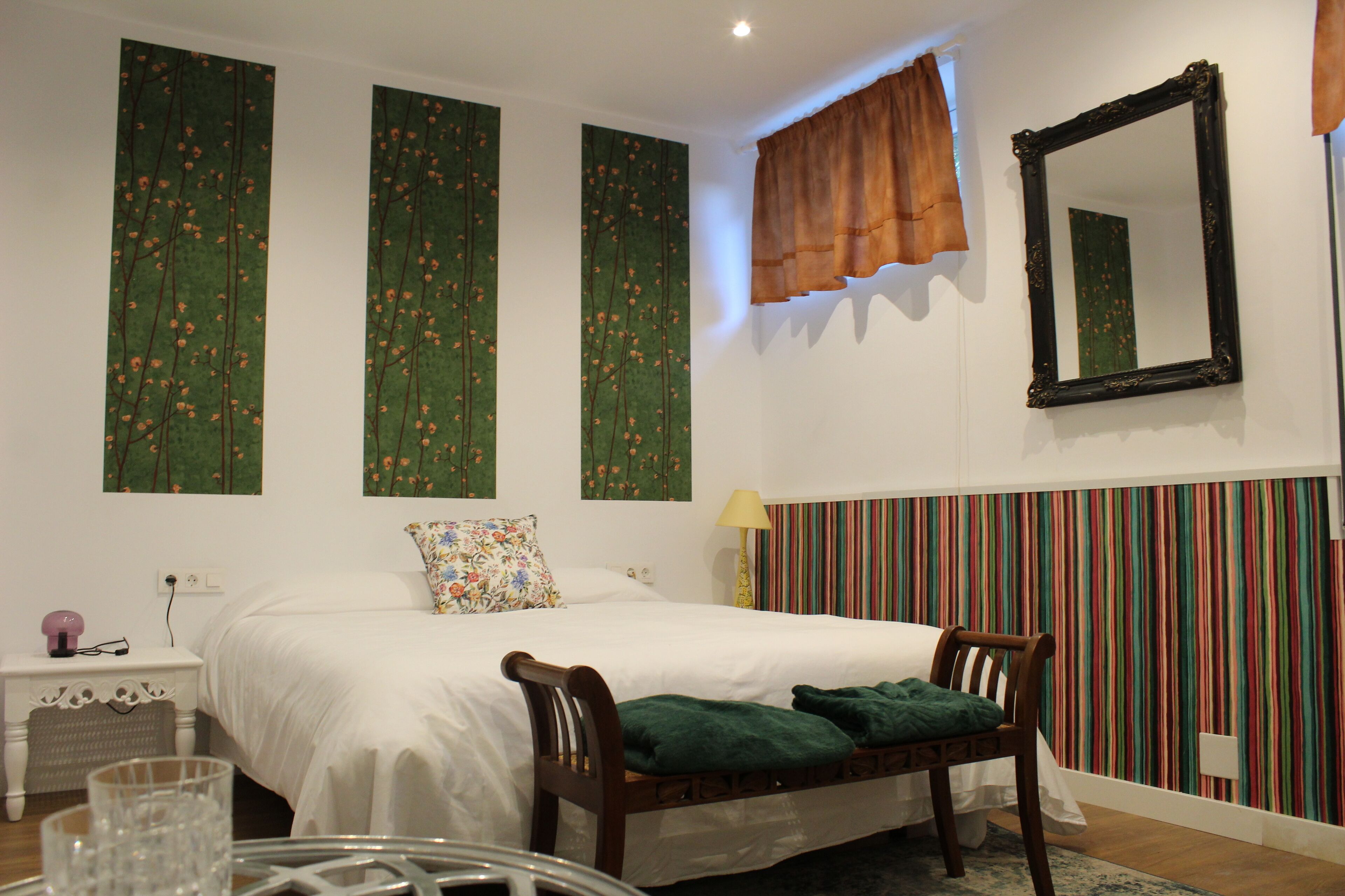 A bedroom with a large bed, a wooden chair, a bedside table, a mirror, and a striped wall panel.