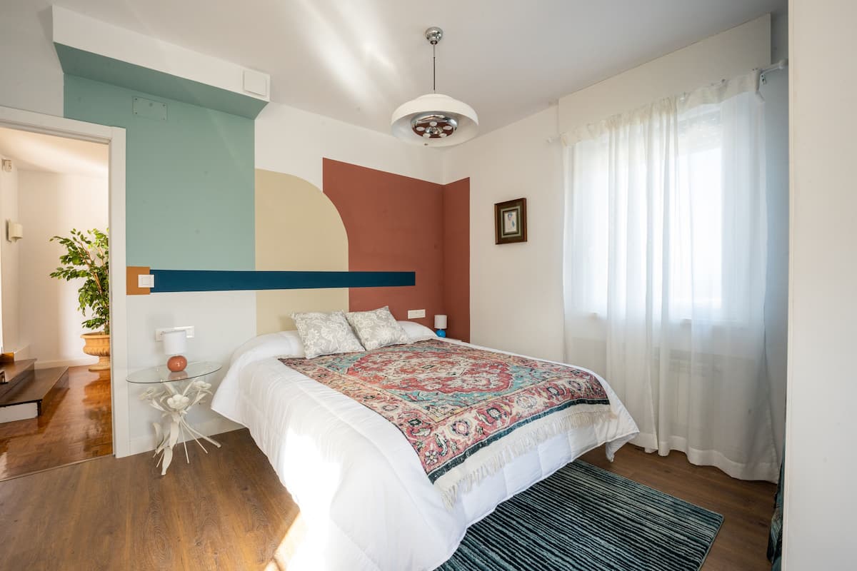 A bedroom with a large bed, a bedside table, a potted plant, and a view of the outside through a window with curtains.