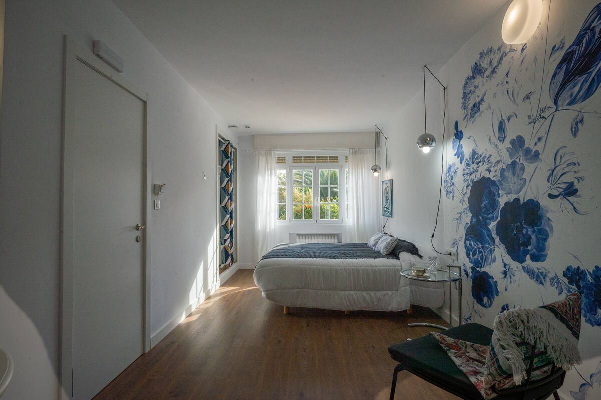 A bedroom with a bed, a bedside table, a chair, a window with curtains, and a floral wall mural.