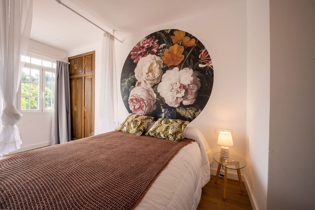 A bedroom with a large floral artwork on the wall, a bed with a brown blanket, and a bedside lamp.