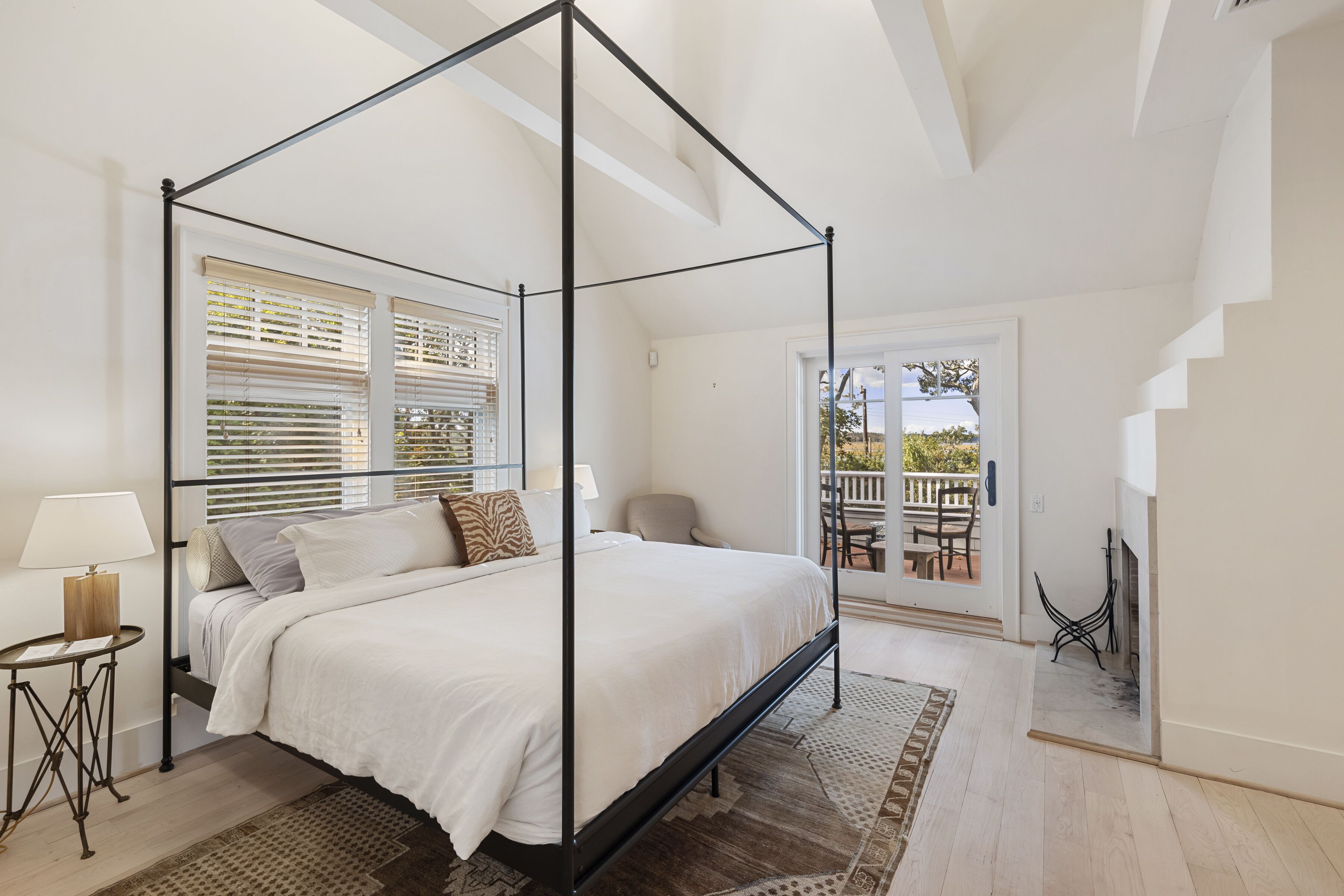 Un lit à baldaquin avec une couvre-lit blanche, une petite table de chevet avec une lampe, et une vue sur l’extérieur par une fenêtre munie de stores.