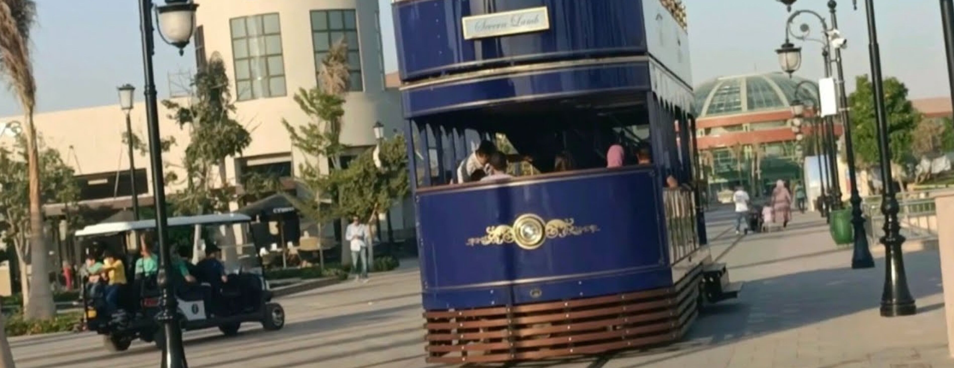 A double-decker tram with passengers, moving along a street lined with palm trees and buildings.