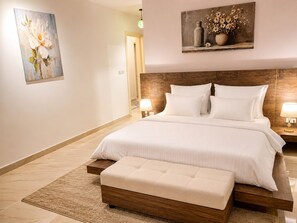 A hotel room with a large bed, a wooden headboard, a bedside table with a lamp, and a painting on the wall. - Casa De Elegant (New Cairo)