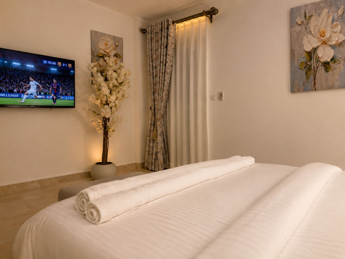 Une chambre d’hôtel avec un lit, un téléviseur diffusant un événement sportif, un vase rempli de fleurs et un tableau accroché au mur.