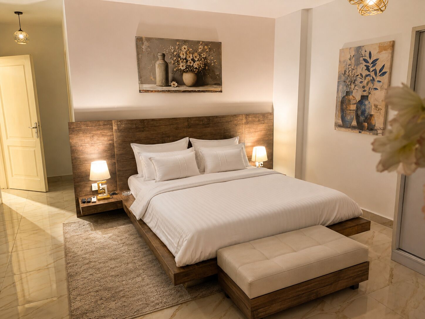 A modern bedroom with a wooden headboard, a large bed with white bedding, two bedside lamps, a nightstand, and a small bedside table.