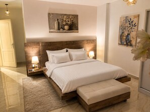 A modern bedroom with a wooden headboard, a large bed with white bedding, two bedside lamps, a nightstand, and a small bedside table. - Casa De Elegant (New Cairo)