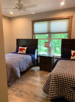 A bedroom with two beds, each with checkered bedding and a pillow with a red bear design. There is a ceiling fan, a window with blinds, and a nightstand between the beds.