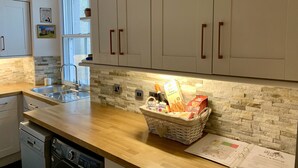 Una cucina con mobili bianchi, zoccolo in pietra, piano di lavoro in legno, lavello, lavastoviglie e un cesto con pane e pasta.